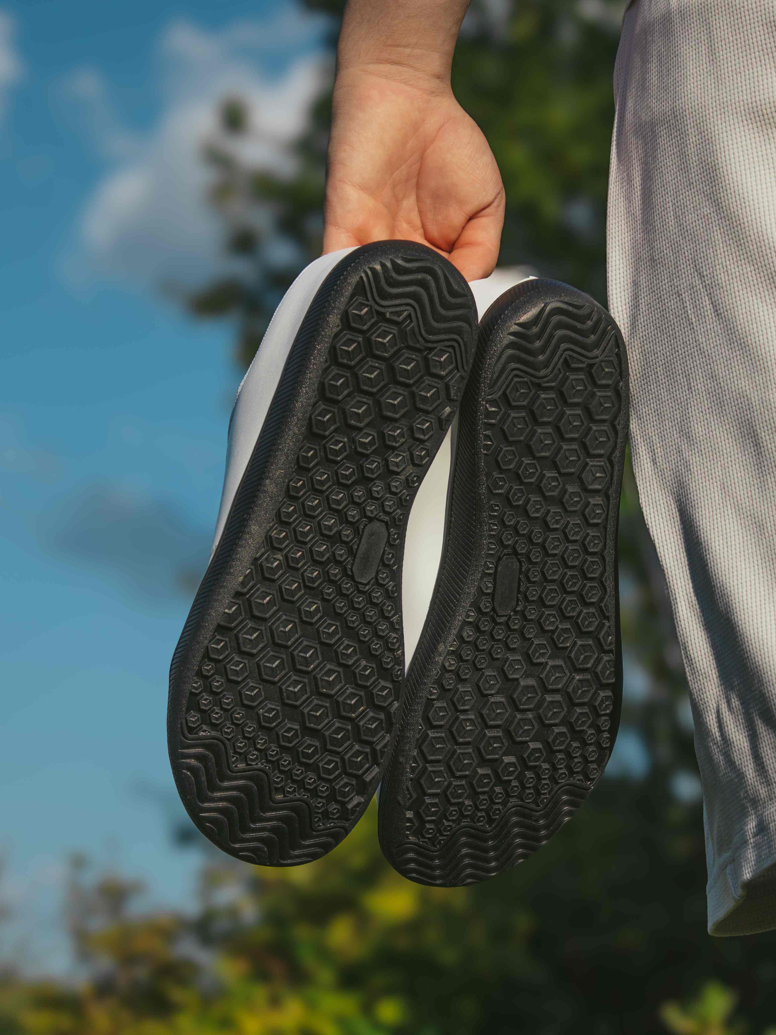 Hand hält ein Paar weiße Grounding-Barfußschuhe Mitochondriak® so, dass die schwarzen Sohlen mit Wabenprofil zu sehen sind